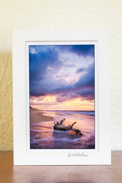 Sunrise drama on a Frisco beach — stormy skies building above the horizon as seafoam squirrels around the base of driftwood, caught in the quiet of the morning tide.
