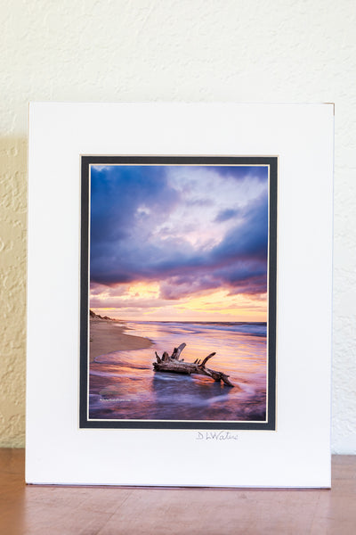 Sunrise drama on a Frisco beach — stormy skies building above the horizon as seafoam squirrels around the base of driftwood, caught in the quiet of the morning tide.