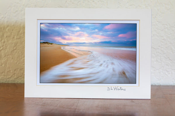 Sunrise with sweping wave at Cape Hatteras National Seashore on the Outer Banks, NC. While watching the waves ebb and flow I envisioned this photo. I knewI wanted the empty beach with the spectacular early morning light on the clouds as the sea foam swept back out to sea with a long enough shutter speed to show the motion.