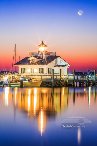 Roanoke Marshes Lighthouse in Manteo North Carolina with the moon setting behind it.   The sun is just brightening the sky and reflecting in the calm waters of Shallowbag Bay.
